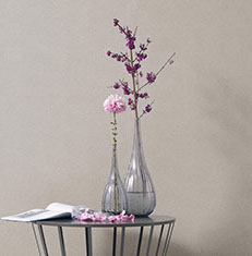 Deux vases en verre posés sur une table ronde métalique devant un mur recouvert d'un tissu beige clair en tenture murale forment une déco zen